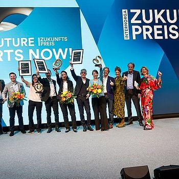 Gewinnerinnen und Gewinner des INTERNORGA Zukunftspreises 2024 jubeln auf der Bühne mit Trophäen, Urkunden und Blumen. Im Hintergrund die Leinwand mit „The Future Starts Now!“