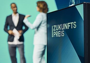 Im Vordergrund ist ein Rednerpult mit der Aufschrift "Zukunftspreis" zu sehen, während im Hintergrund unscharf eine Frau in einem hellen Anzug und ein Mann in dunkler Kleidung auf der Bühne miteinander sprechen.