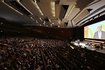 Szene beim Foodservice Forum. Großer Konferenzsaal mit vollem Publikum, zwei Rednern auf der Bühne und einer Leinwand mit einem Rednerbild.