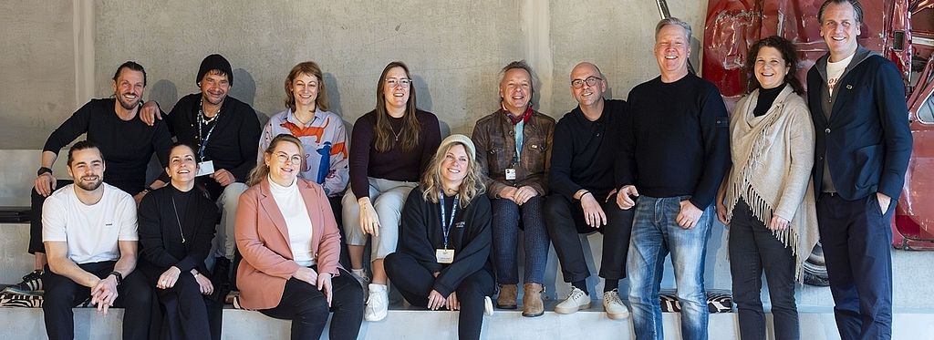 Eine Gruppe von 13 Personen sitzt entspannt auf Stufen in einem modern gestalteten Raum. Sie lächeln in die Kamera, im Hintergrund ist ein in die Wand integriertes, rotes Oldtimer-Frontteil zu sehen.