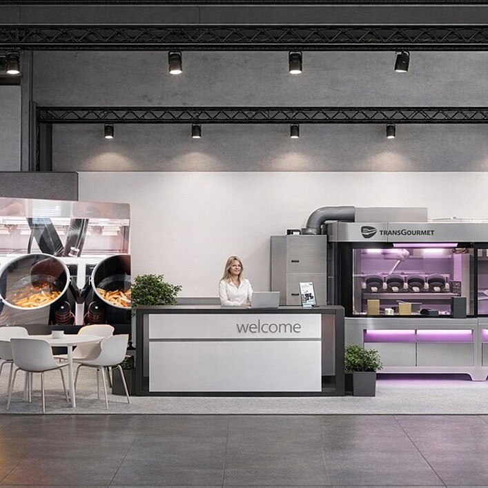 Messestand in modernem, offenen Design. In der Mitte steht ein Empfangstresen mit der Aufschrift „welcome“, hinter dem eine Person steht. Links befinden sich mehrere kleine Tische mit weißen Stühlen sowie eine große Wand mit einem Küchenmotiv. Rechts sind professionelle Gastronomiegeräte und Automaten mit lila Beleuchtung zu sehen, darüber das Logo „food@service“. Daneben stehen zwei blaue Informations- oder Bestellterminals.