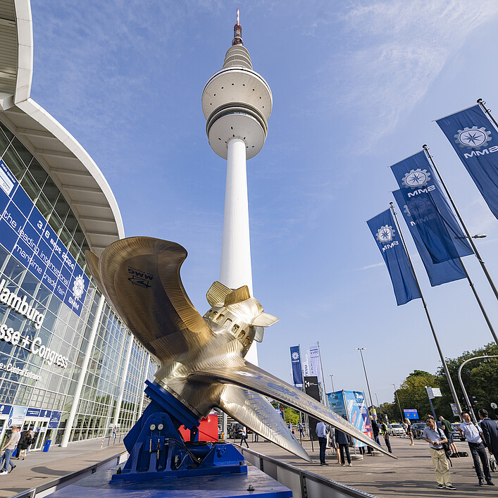View of the Hamburg exhibition grounds with the Heinrich Hertz Tower in the center. A large ship propeller sculpture is displayed in the foreground, while several blue trade fair flags are flying on the right. Visitors are walking near the Hamburg Messe & Congress entrance under a clear blue sky.