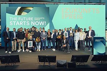 Eine große Gruppe von Menschen, darunter Preisträger und Veranstalter, posiert auf einer Bühne mit Trophäen und Urkunden vor einer Leinwand mit der Aufschrift "Zukunftspreis" und "The Future Starts Now!".