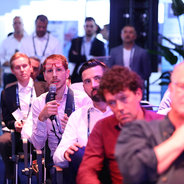 A person is seated in the audience at an event, holding a microphone while asking a question or making a comment. The setting appears to be a busy conference or networking space with several attendees seated and standing in the background, listening attentively.