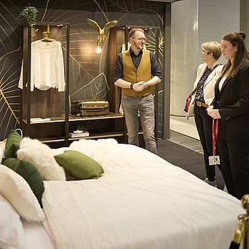 Eine stilvolle Schlafzimmerausstellung mit einem gemütlich dekorierten Bett in Weiß und Grüntönen im Vordergrund. Im Hintergrund steht ein Mann in einer gelben Weste und unterhält sich mit zwei Frauen, die das Design begutachten.