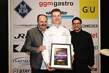 Der Gewinner des Next Chef Awards 2025, Gedion Beckmann, hält stolz seine Siegerurkunde, während Sternekoch Johann Lafer (links) und Moderator Sebastian E. Merget (rechts) ihn gratulierend flankieren.