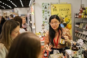 Eine Standmitarbeiterin von „Dr. Jaglas“ erklärt Besucherinnen ein Produkt. Sie hält eine Flasche in der Hand und gestikuliert, während sich auf dem Stand verschiedene Kräuterliköre und andere Spezialitäten befinden.