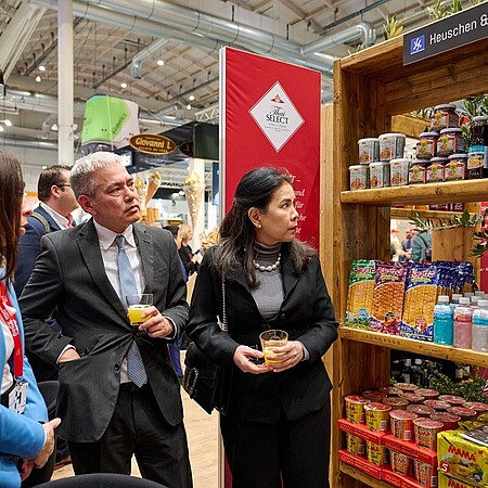 Mehrere Personen stehen auf einer Messe vor einem Ausstellungsregal mit Lebensmitteln und Getränken des Unternehmens Heuschen & Schroo und unterhalten sich, während sie Getränke in der Hand halten.
