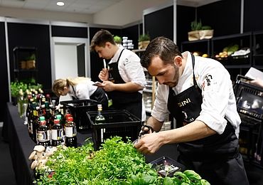 Drei Köche arbeiten während des Next Chef Awards an einer Küchenstation mit frischen Kräutern und Zutaten. Ein Koch schneidet Kräuter im Vordergrund.