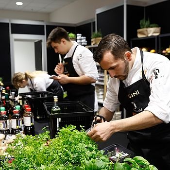 Drei Köche arbeiten während des Next Chef Awards an einer Küchenstation mit frischen Kräutern und Zutaten. Ein Koch schneidet Kräuter im Vordergrund.