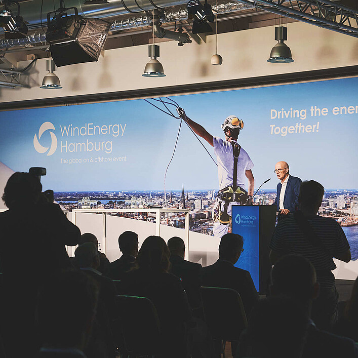Person spricht an einem Rednerpult vor Publikum auf einer Bühne der WindEnergy Hamburg. Im Hintergrund ist eine großformatige Veranstaltungswand mit WindEnergy-Hamburg-Logo und Hamburg-Panorama zu sehen.