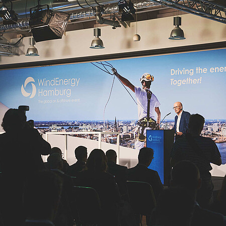 Person spricht an einem Rednerpult vor Publikum auf einer Bühne der WindEnergy Hamburg. Im Hintergrund ist eine großformatige Veranstaltungswand mit WindEnergy-Hamburg-Logo und Hamburg-Panorama zu sehen.