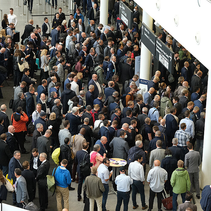 Eine große Menschenmenge steht in einem belebten Eingangsbereich einer Messe. Die Leute unterhalten sich und bewegen sich durch den Raum.