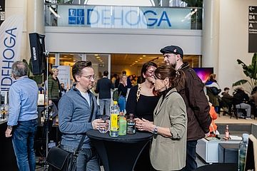 Eine Gruppe von Menschen steht an einem Stehtisch bei einer DEHOGA-Veranstaltung und unterhält sich. Im Hintergrund sind das DEHOGA-Logo und weitere Messebesucher zu sehen. Auf dem Tisch stehen verschiedene Getränke.