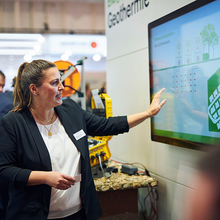 Eine Frau präsentiert auf einer Messe eine große digitale Anzeige zum Thema Geothermie. Sie zeigt auf ein Diagramm, während mehrere Personen aufmerksam zuhören.