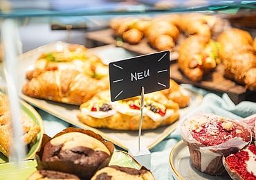 Nahaufnahme einer Auslage in einer Bäckerei mit verschiedenen Backwaren wie Croissants und Muffins. Ein kleines Schild mit der Aufschrift „NEU“ hebt neue Produkte hervor.