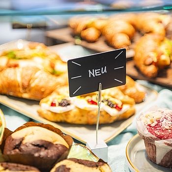 Nahaufnahme einer Auslage in einer Bäckerei mit verschiedenen Backwaren wie Croissants und Muffins. Ein kleines Schild mit der Aufschrift „NEU“ hebt neue Produkte hervor.