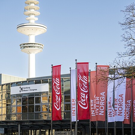 Außenansicht des Hamburg Messe und Congress Gebäudes bei klarem Himmel. Vor dem Eingang stehen mehrere rote und weiße Fahnen mit Coca-Cola- und INTERNORGA‑Branding. Im Hintergrund ragt der markante Heinrich-Hertz-Turm in den Himmel.