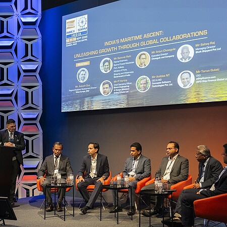 Eine Podiumsdiskussion auf einer Bühne mit sieben Personen, von denen eine am Rednerpult steht und sechs in einer Reihe auf orangefarbenen Sesseln sitzen. Hinter ihnen zeigt eine große Leinwand den Titel der Veranstaltung zum Thema maritime Entwicklung und internationale Zusammenarbeit. Die Bühne ist modern ausgeleuchtet und hat eine geometrische Wandgestaltung. Das Publikum ist nicht sichtbar.