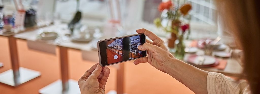 Eine Person fotografiert mit einem Smartphone den „Skywalk“, eine modern gestaltete Fläche mit elegant gedeckten Tischen, floralen Dekorationen und großen Fenstern, die für eine lichtdurchflutete Atmosphäre sorgen.