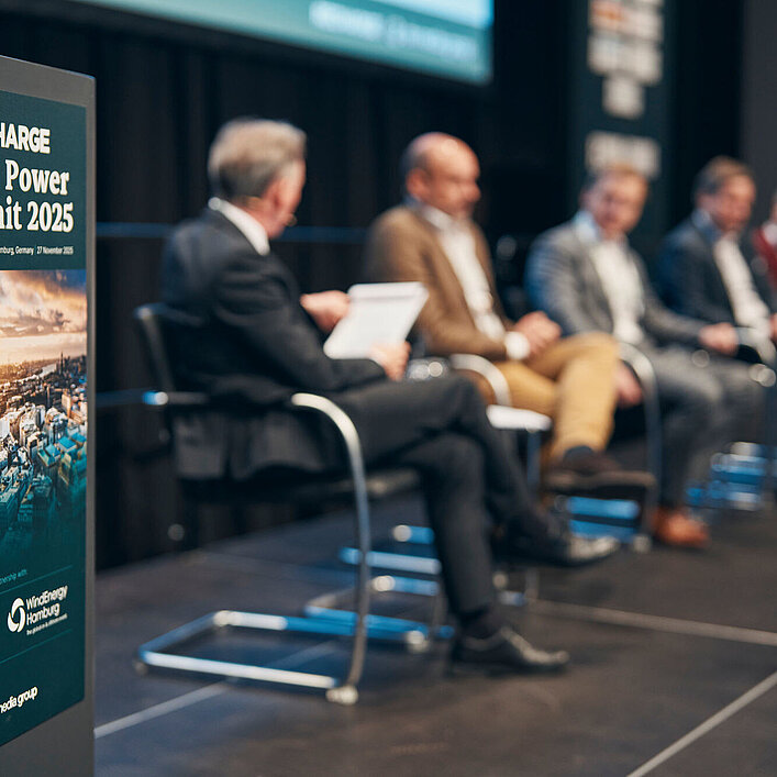 Podiumsdiskussion beim „Wind Power Summit 2025“ mit mehreren Fachleuten auf der Bühne, Fokus auf Veranstaltungsplakat im Vordergrund.